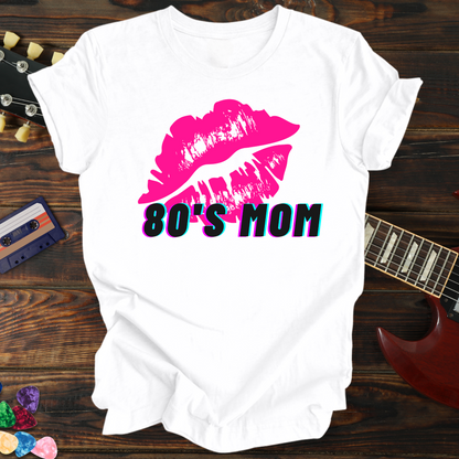 White t-shirt with pink lips and '80's Mom' text on a wooden surface with a guitar and cassette tape.
